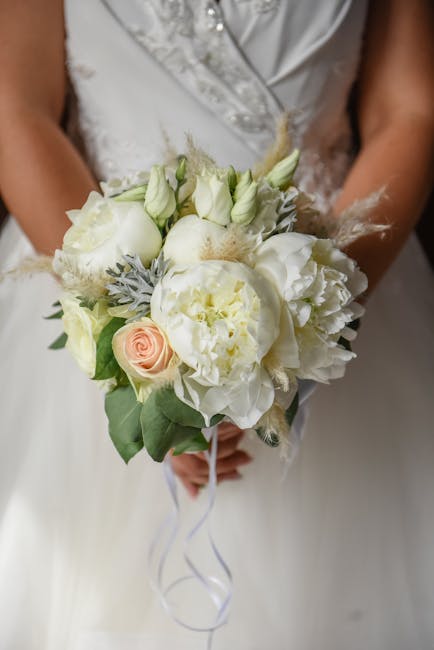 A bridal bouquet held by a bride dressed in a white wedding gown with lace and satin details. The bouquet features a delicate arrangement of fresh flowers, including large white peonies, soft cream ro