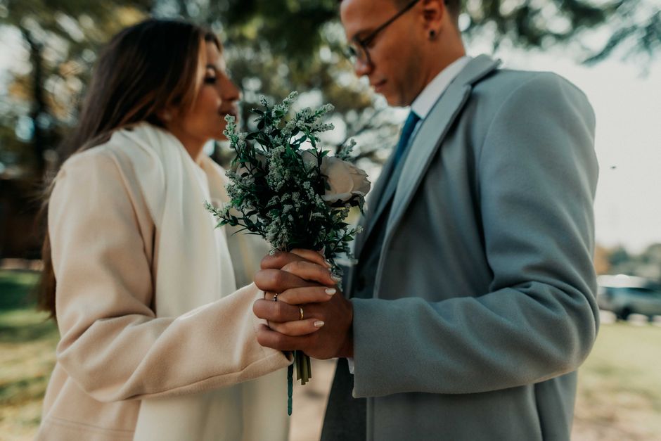 Close-up of a bride and groom holding a floral bouquet together during a wedding in an outdoor park setting with green trees in the background. The bouquet features a mix of white roses, small white f