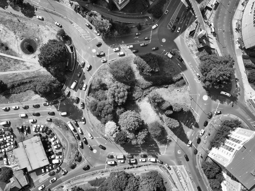 A black and white aerial view of a busy roundabout surrounded by trees, cars, and buildings, with a small park area in the center containing a mix of leafy trees and grassy patches. The traffic is spr