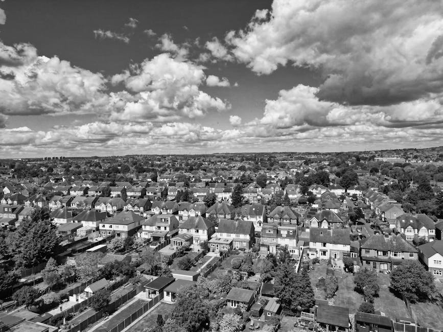 A black and white aerial photograph of a residential suburban area, showing rows of detached and semi-detached houses with pitched roofs, neatly arranged gardens, and tree-lined streets. The sky above